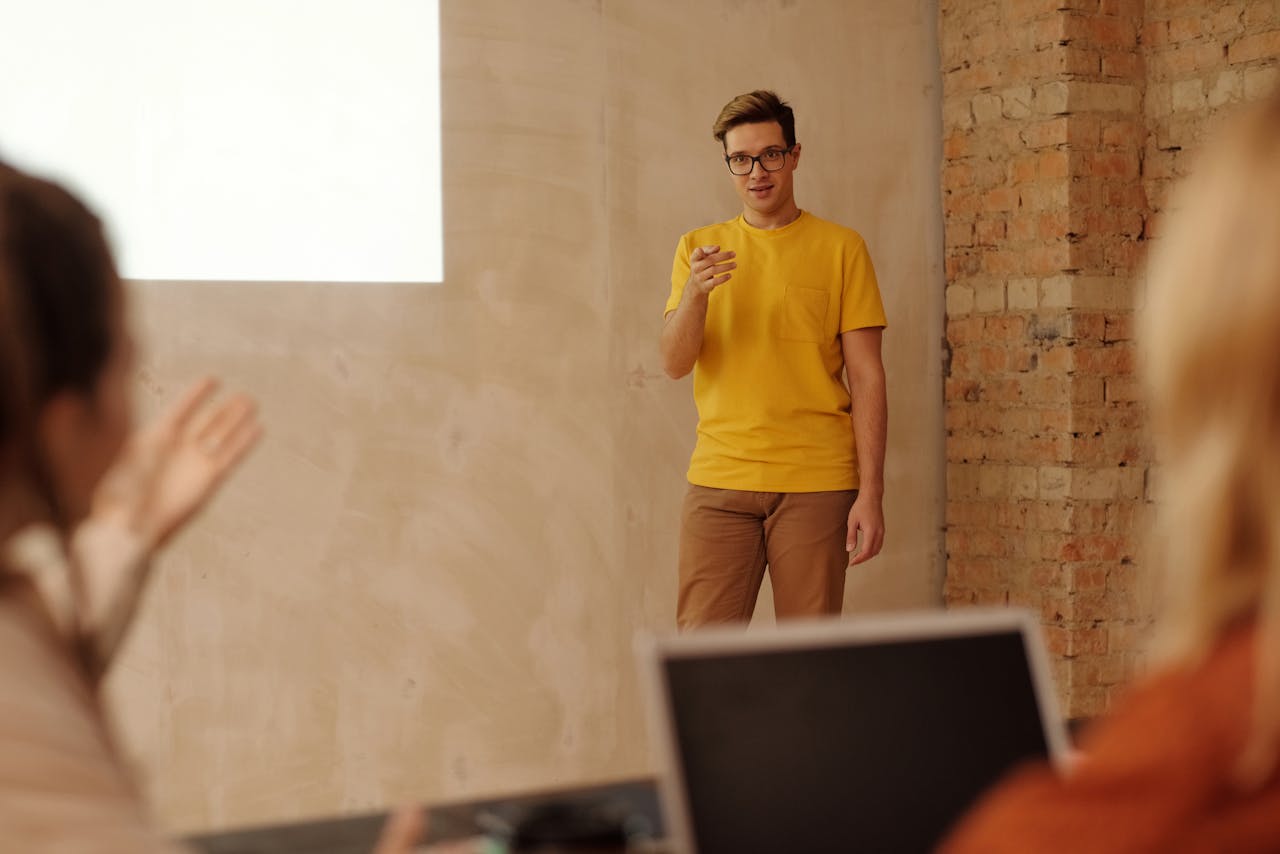 A young man in a yellow shirt presents information to colleagues in a casual meeting setting.